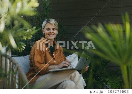 Woman in the garden working on laptop, sitting on patio chair. Businesswoman working remotely from outdoor home office, thinking about new business or creative idea. Concept of green outdoor home 108272608