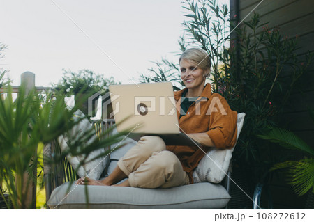 Woman in the garden working on laptop, sitting on patio chair. Businesswoman working remotely from outdoor home office, thinking about new business or creative idea. Concept of green outdoor home 108272612