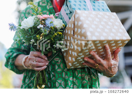 Close up of senior birthday woman holding gifts and bouquet in hands. Garden birthday party for the senior lady. Close up of senior birthday woman holding gifts and bouquet in hands. Garden birthday party for the senior lady. 108272640
