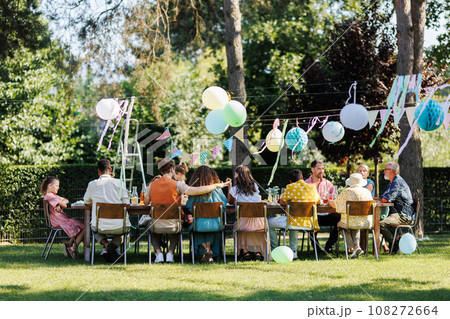Family and friends sitting at the party table during a summer garden party outdoors. Rear view of people sitting at the table. 108272664
