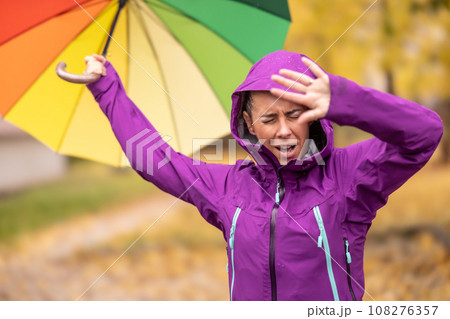 A young woman with an umbrella in a rain jacket wears a hood and protects her head from rain and strong wind during late autumn A young woman with an umbrella in a rain jacket wears a hood and protects her head from rain and strong wind during late autumn 108276357