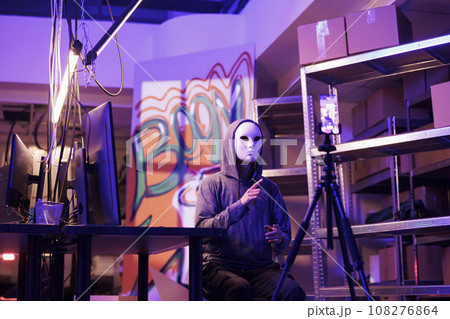 Hacker in anonymous mask threatening online while streaming live in abandoned warehouse. Internet scammer recording video with ransom on smartphone in hideout place at night 108276864