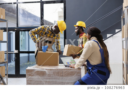 African american warehouse managers team preparing customer order for delivery. All black storehouse employees packing cardboard box with sticky tape and checking goods list on laptop African american warehouse managers team preparing customer order for delivery. All black storehouse employees packing cardboard box with sticky tape and checking goods list on laptop 108276963