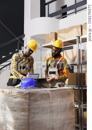 Logistics managers analyzing inventory and comparing data on laptop and clipboard in warehouse. African american men checking goods supply schedule and working in distribution department storehouse 108276975