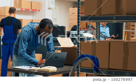 Small business owner planning supplies shipment at storehouse, putting merchandise in cardboard boxes for distribution. Man taking goods from storage shelves to ship products. Handheld shot. Small business owner planning supplies shipment at storehouse, putting merchandise in cardboard boxes for distribution. Man taking goods from storage shelves to ship products. Handheld shot. 108276997