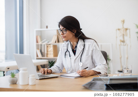 Female doctor with laptop writing prescription at office Female doctor with laptop writing prescription at office 108277905