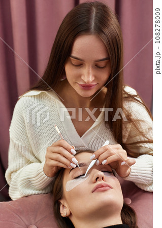 professional beautician with tweezers makes eyelash extension for beautiful woman in beauty salon. 108278009