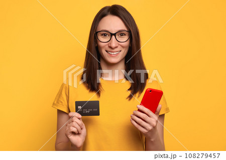 Satisfied pretty girl in glasses holding mobile phone and credit card isolated on yellow wall 108279457