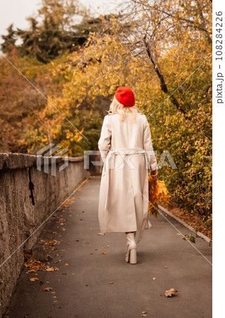 autumn woman in a red beret, a light coat and a red skirt, against the backdrop of an autumn park with yellow leaves in her hands autumn woman in a red beret, a light coat and a red skirt, against the backdrop of an autumn park with yellow leaves in her hands 108284226