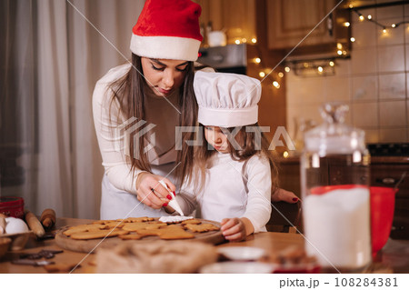 Mom in santas hat with her little doughter decorates gingerbread at home. Christmas and New Year traditions concept. Christmas bakery. Happy holidays Mom in santas hat with her little doughter decorates gingerbread at home. Christmas and New Year traditions concept. Christmas bakery. Happy holidays 108284381