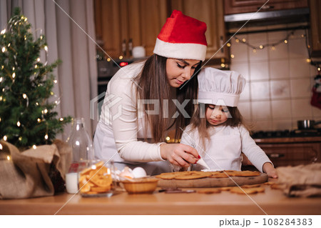 Happy little girl with her beautiful mom make gingerbread at home. Christmas decoration at kitchen. Fir tree with fairy lights Happy little girl with her beautiful mom make gingerbread at home. Christmas decoration at kitchen. Fir tree with fairy lights 108284383