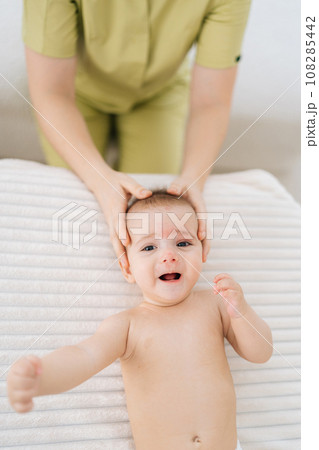 Vertical top view of adorable infant baby boy being manipulated by unrecognizable osteopathic manual therapist or physician. Closeup hands of female masseuse massage head of small newborn kid. Vertical top view of adorable infant baby boy being manipulated by unrecognizable osteopathic manual therapist or physician. Closeup hands of female masseuse massage head of small newborn kid. 108285442