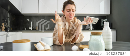 Attractive young cheerful girl baking at the kitchen, making dough, holding recipe book, having ideas 108287084