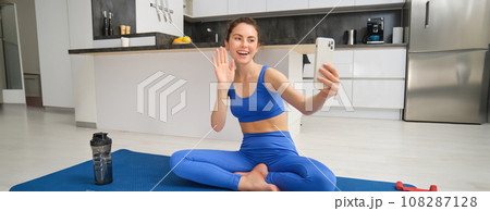 Vlogger does sports, records her workout training from home on smartphone camera, posing for selfie inside her house, sits on rubber yoga mat in blue leggings and sportsbra Vlogger does sports, records her workout training from home on smartphone camera, posing for selfie inside her house, sits on rubber yoga mat in blue leggings and sportsbra 108287128