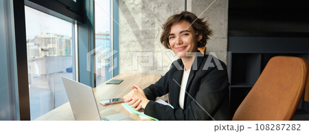 Portrait of working woman in an office, using laptop and mobile phone, sitting near the window in coworking space 108287282