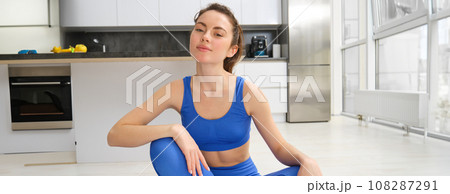 Portrait of young, healthy and fit brunette woman, sits on rubber mat, does yoga, workout training at home, wears blue fitness sportswear and smiles 108287291