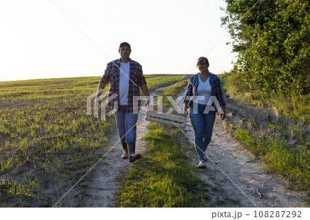 A couple of farmers carry a box of potatoes across the field against the backdrop of a beautiful sunset A man and a woman together carry a heavy box of vegetables harvested and grown in their fields. 108287292