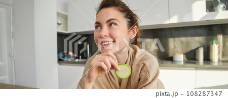 Portrait of happy, smiling young woman in the kitchen, cooking, chopping zucchini, holding vegetables and looking happy, preparing vegan food meal at home 108287347