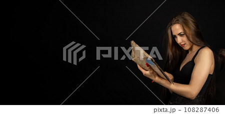 Halloween. Portrait. A beautiful woman with black makeup holds an ancient spell book in her hands on a black background. The witch is studying an old book holding it in her hands. Copy space. 108287406