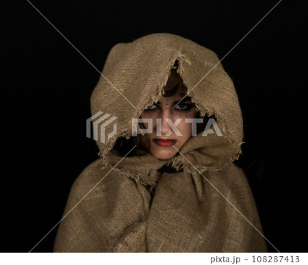 In a cape with a hood, a beautiful female witch stands in the semi-darkness with runic symbols on her face. Halloween. Halloween black witch portrait. Halloween holiday concept. 108287413