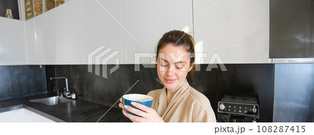Portrait of smiling woman drinks coffee, stands in the kitchen and enjoys delicious cup of cappuccino in the morning, looks happy at camera Portrait of smiling woman drinks coffee, stands in the kitchen and enjoys delicious cup of cappuccino in the morning, looks happy at camera 108287415