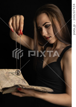 On Halloween, the witch performs a ritual with a book and a talisman. A beautiful female witch reads fortunes from an ancient book, holding a talisman in her hand. Halloween divination concept. 108287434