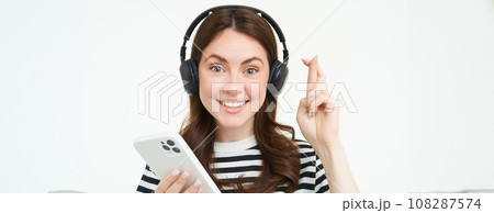 Young woman holding fingers crossed, making wish, hoping for something, holding smartphone, wearing wireless headphones, white background 108287574
