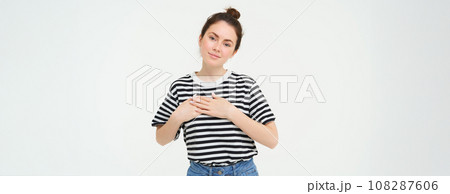 Portrait of happy, beautiful brunette woman, holds hands on heart and smiles, stands in casual clothes over white background Portrait of happy, beautiful brunette woman, holds hands on heart and smiles, stands in casual clothes over white background 108287606