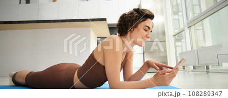 Indoor shot of fit and healthy woman workout at home, lying on yoga mat while using smartphone, watching fitness training at mobile phone, resting after exercises 108289347