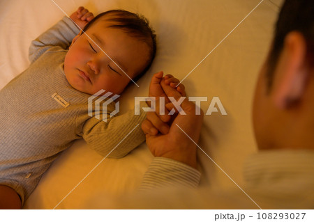 赤ちゃんをベッドに寝かせる父親　指を握る子ども　イクメン　寝かしつけ　添い寝　把握反射　0歳乳幼児 108293027