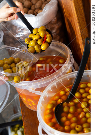 Hands of buyer pouring pickled olives by weight into plastic container Hands of buyer pouring pickled olives by weight into plastic container 108293391