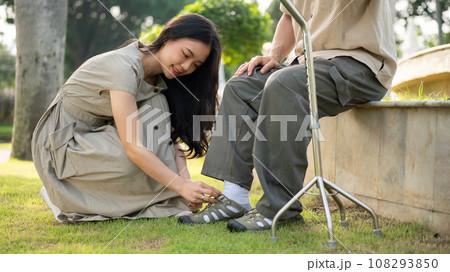 A caring Asian daughter is helping her senior dad put shoes on while relaxing in a park together. A caring Asian daughter is helping her senior dad put shoes on while relaxing in a park together. 108293850