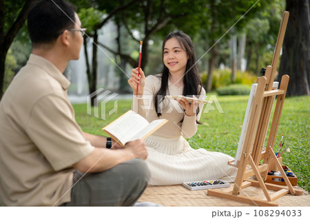 A lovely Asian girl enjoys painting with her dad in a park, picnicking, and spending time together. 108294033