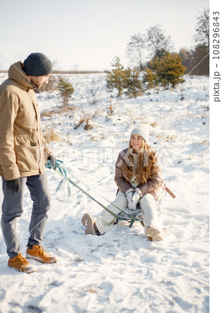 Portrait of romantic couple having fun on winter walk. Man carry his wife on a sled. Family wearing warm jacket and knitted hats. 108296843