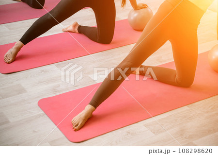 A group of six athletic women doing pilates or yoga on pink mats in front of a window in a beige loft studio interior. Teamwork, good mood and healthy lifestyle concept. 108298620