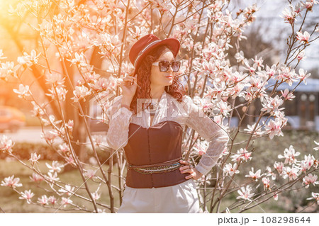 Magnolia park woman. Stylish woman in a hat stands near the magnolia bush in the park. Dressed in white corset pants and posing for the camera. 108298644