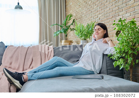 Relaxed young woman with raised arms sitting on sofa in living room Relaxed young woman with raised arms sitting on sofa in living room 108302072