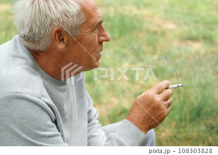 Senior man lighting up a cigarette outdoors against green nature background. Old thoughtful man smoking, looking aside. Close Up Face. Relaxed pensioner enjoy weekend rest outside. Unhealthy habits Senior man lighting up a cigarette outdoors against green nature background. Old thoughtful man smoking, looking aside. Close Up Face. Relaxed pensioner enjoy weekend rest outside. Unhealthy habits 108303828
