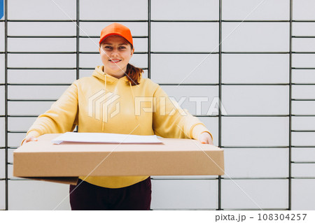 Happy 30s delivery woman in red hat giving packages. Concept of fast delivery to automatic self lockers 108304257