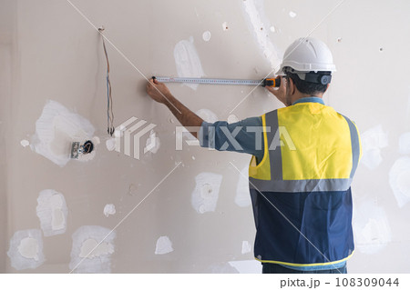 Skilled worker standing near shabby wall measuring distance with tape-line before drilling holes man in protective hardhat and vast renovates living room interior Skilled worker standing near shabby wall measuring distance with tape-line before drilling holes man in protective hardhat and vast renovates living room interior 108309044