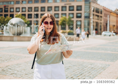 Beautiful smiling female with paper map talking on Phone and standing outdoors. Phone Communication. Happy cheerful young woman walking on city street, Urban lifestyle concept. Traveler Beautiful smiling female with paper map talking on Phone and standing outdoors. Phone Communication. Happy cheerful young woman walking on city street, Urban lifestyle concept. Traveler 108309362