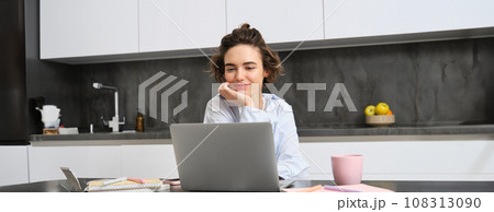 Young female entrepreneur works from homes. Girl with laptop sits in her kitchen, does homework, works on remote, connects to online meeting, taking notes Young female entrepreneur works from homes. Girl with laptop sits in her kitchen, does homework, works on remote, connects to online meeting, taking notes 108313090
