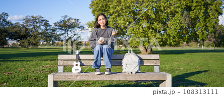 Girl with sad face, sitting on bench with smartphone and ukulele, looking upset and disappointed, being alone in park 108313111