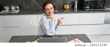 Beautiful woman managing her work from home, drinking coffee and looking at laptop, sitting in kitchen with paperwork, doing her taxes, studying indoors on remote Beautiful woman managing her work from home, drinking coffee and looking at laptop, sitting in kitchen with paperwork, doing her taxes, studying indoors on remote 108313244