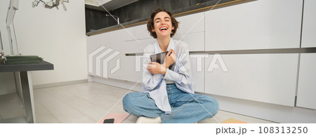 Portrait of happy woman with tablet, sitting on floor, laughing and smiling, working from home on remote, studying online 108313250