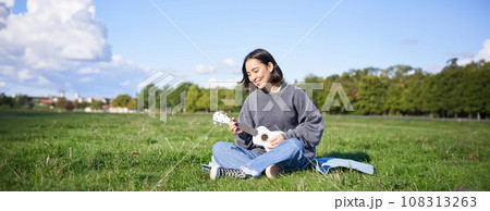 Singing asian girl playing ukulele on grass, sitting on blanket in park, relaxing outdoors on sunny day 108313263