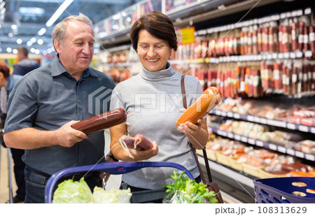 Elderly couple of customers looking for sausage in meat section of supermarket Elderly couple of customers looking for sausage in meat section of supermarket 108313629