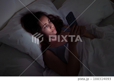 A late-night scene: a woman, bed-bound, captivated by her phone's screen, embodying the challenges of insomnia and smartphone dependency. A late-night scene: a woman, bed-bound, captivated by her phone's screen, embodying the challenges of insomnia and smartphone dependency. 108314093