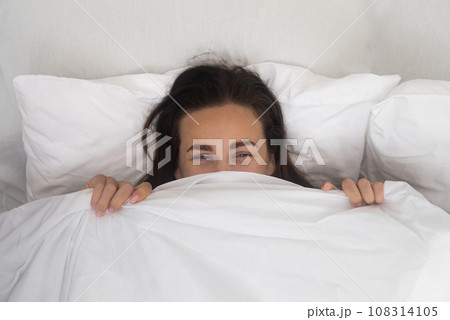 Attractive young woman smiles cheerfully covering face with white blanket positive brunette female feels happiness relaxing in comfortable bed in morning 108314105