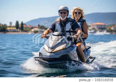Happy senior Caucasian couple in safety helmets and vests riding jet ski on a lake or along sea coast. Active elderly people having fun on water scooter. Healthy lifestyle for retired persons. Happy senior Caucasian couple in safety helmets and vests riding jet ski on a lake or along sea coast. Active elderly people having fun on water scooter. Healthy lifestyle for retired persons. 108316858